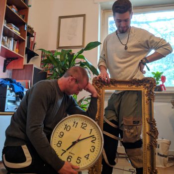 Zwei Moderatoren mit einer Wanduhr und einem Bilderrahmen